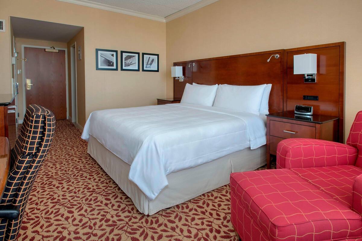 A hotel room with a large bed, a red armchair, a wooden headboard, and a patterned carpet.