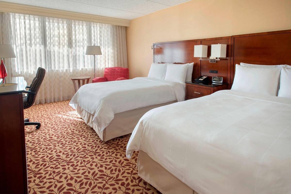 A hotel room with two beds, a desk, a chair, a lamp, and a window with curtains.