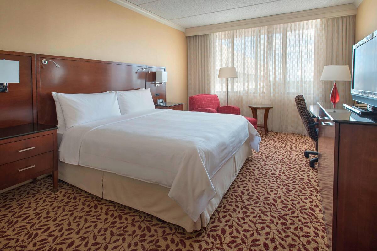 A hotel room with a large bed, a desk with a television, a red chair, and a small table.
