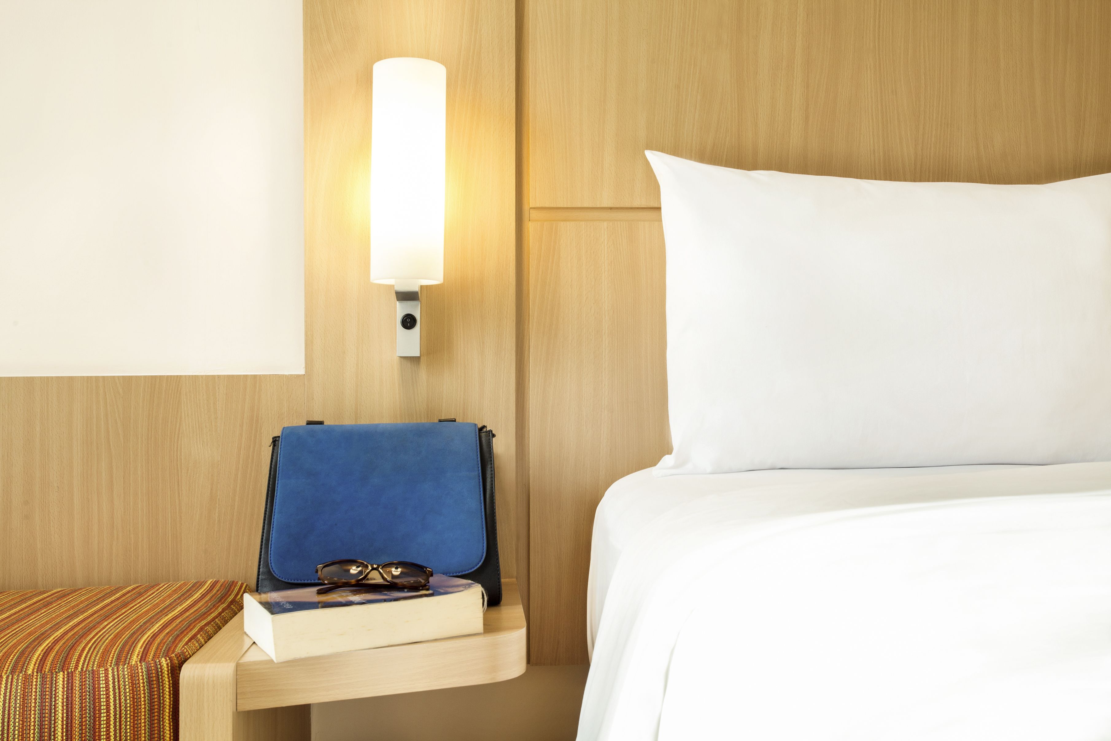 A hotel room with a bed, a bedside table with a lamp, a blue bag, and a book.