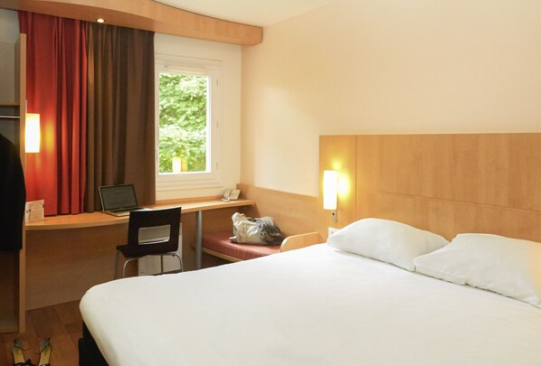 A hotel room with a bed, desk, chair, and a window with a view of greenery. - ibis Sisteron (Sisteron)