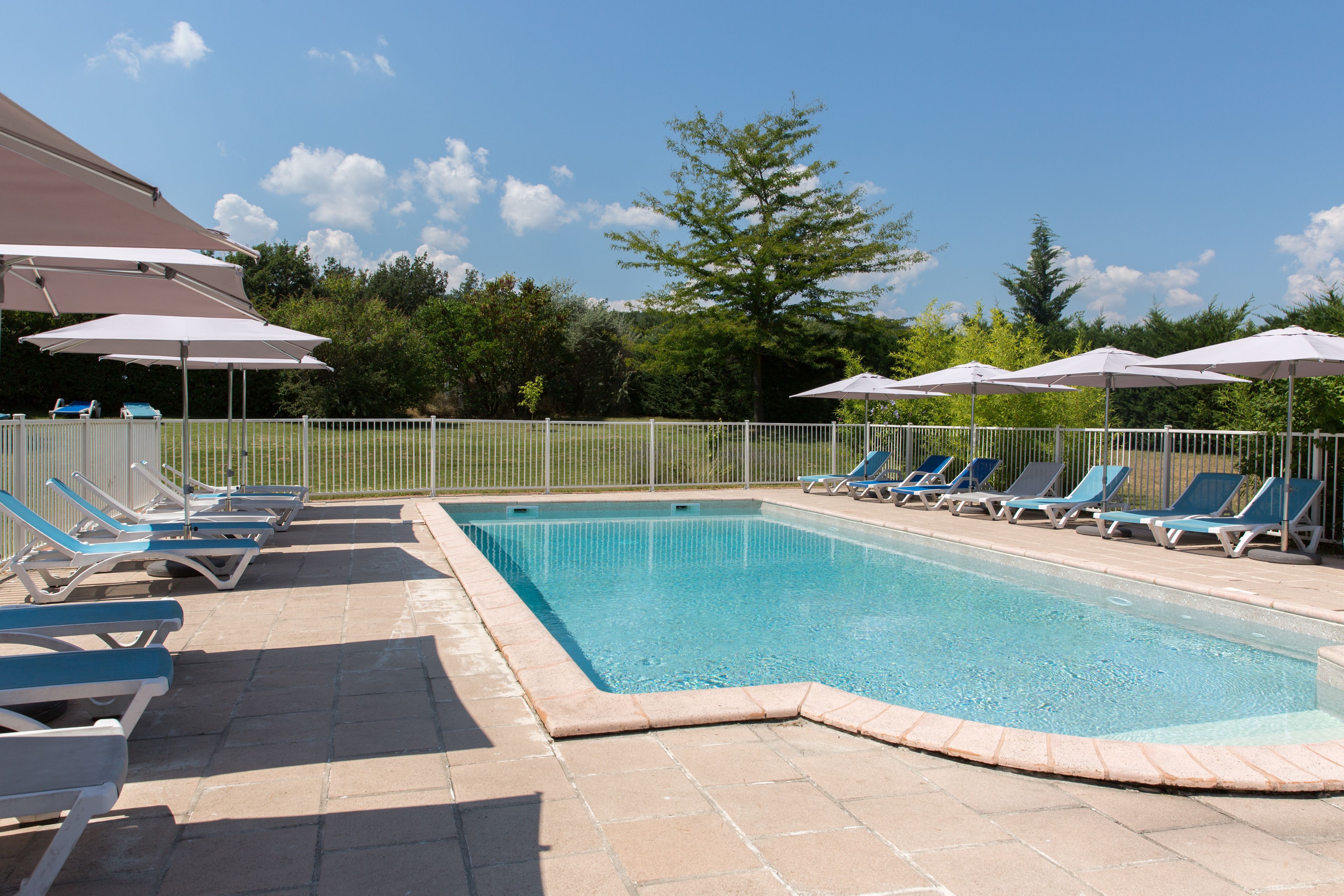 Piscina con sillas de descanso y sombrillas, rodeada de una valla y árboles.