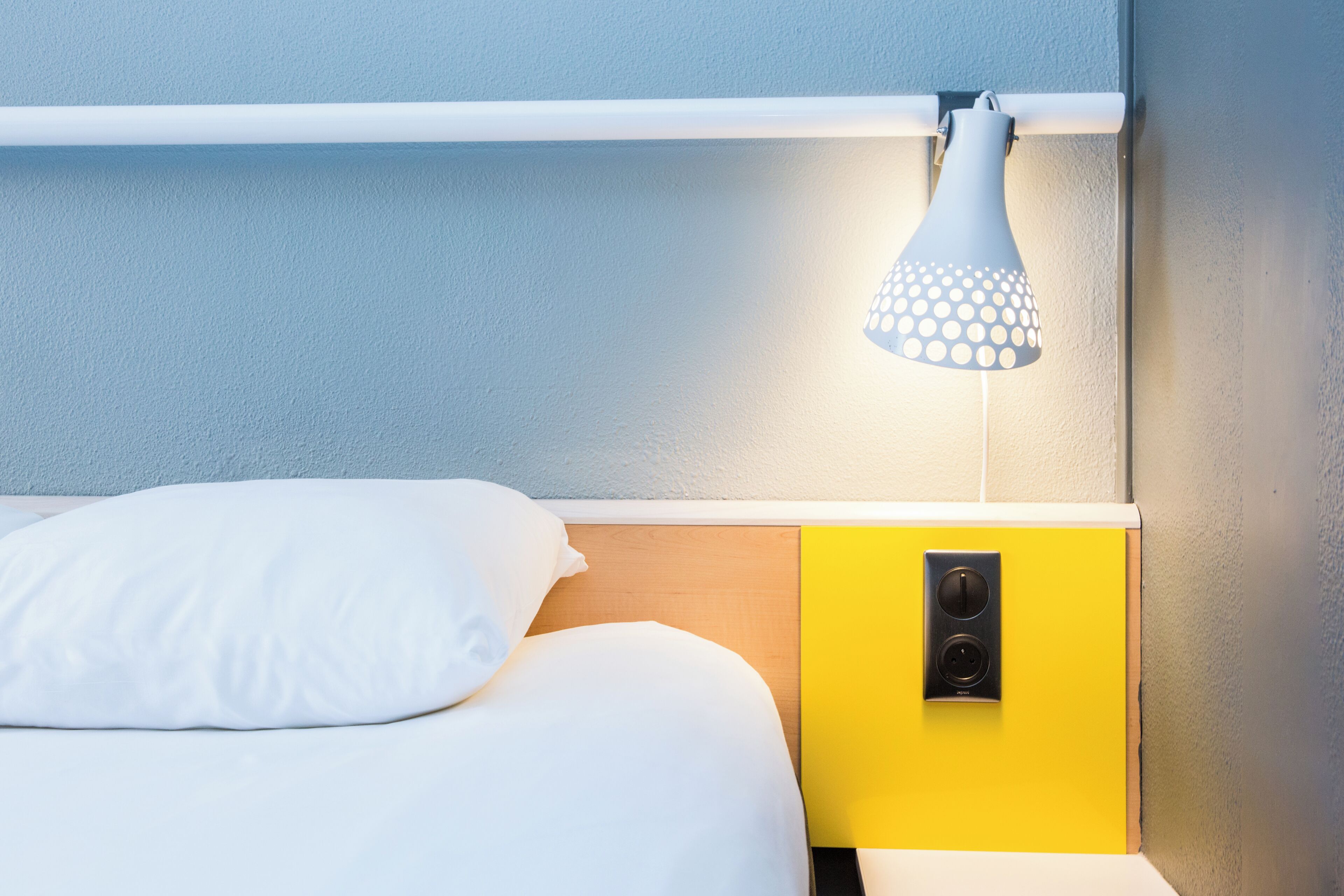 A bed with white bedding, a yellow bedside table with a power outlet, and a white lamp with a circular shade.