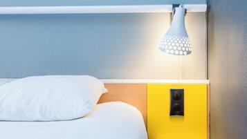 A bed with white bedding, a yellow bedside table with a power outlet, and a white lamp with a circular shade.