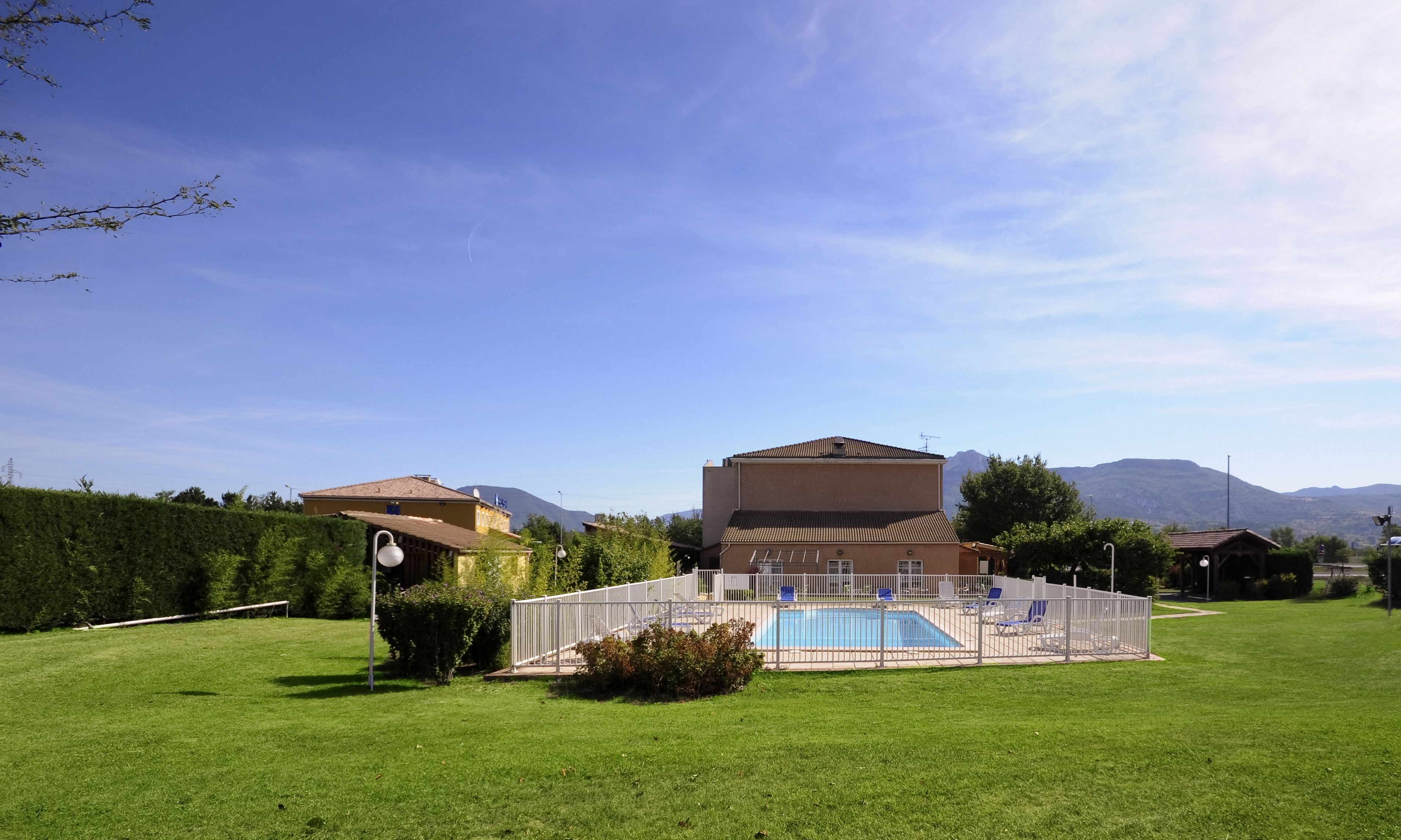 A swimming pool with a fenced area, surrounded by a well-maintained lawn and a building with a tiled roof.