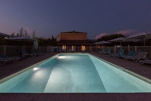 Una piscina con sillas de descanso y sombrillas, rodeada por una cerca y un edificio al fondo.
