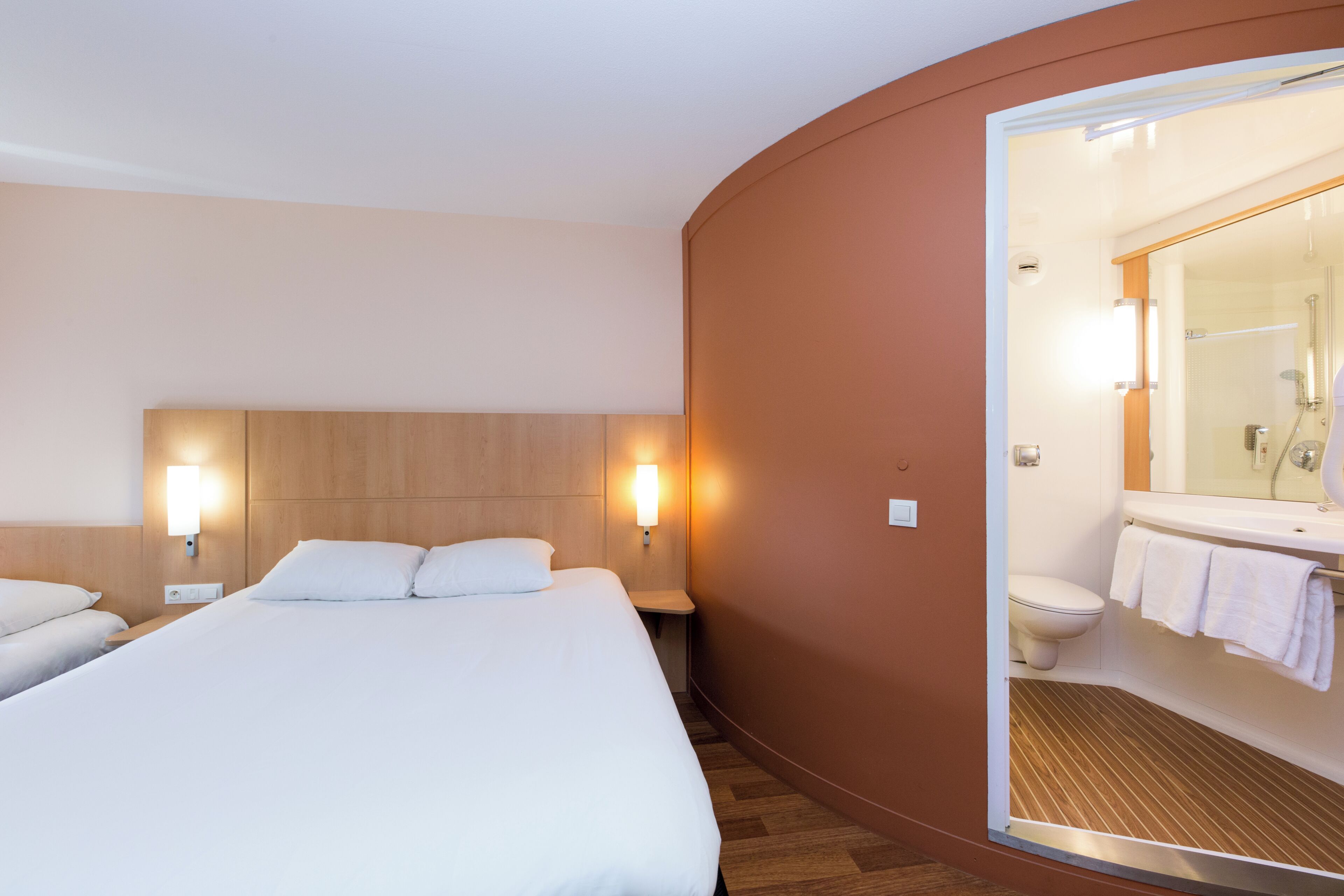 Habitació d'hotel amb un llit gran, dues taules de nit, una làmpada penjada a la paret i un bany amb dutxa i vàter.