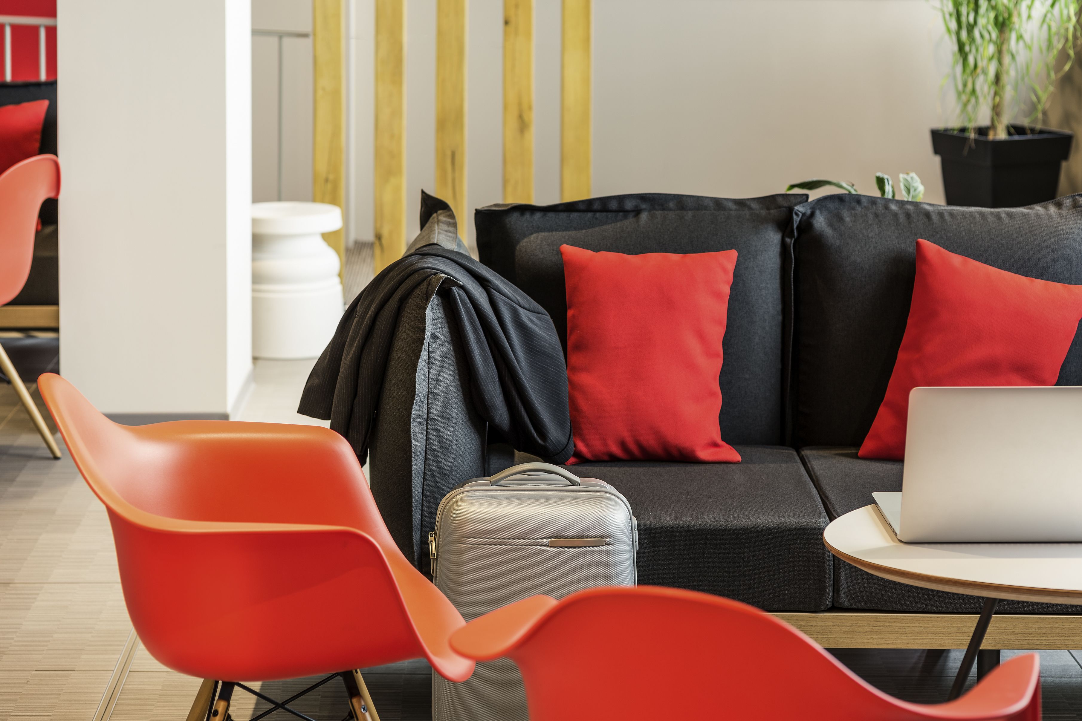 A modern room with a black sofa, red cushions, a grey suitcase, and a laptop on a small table.