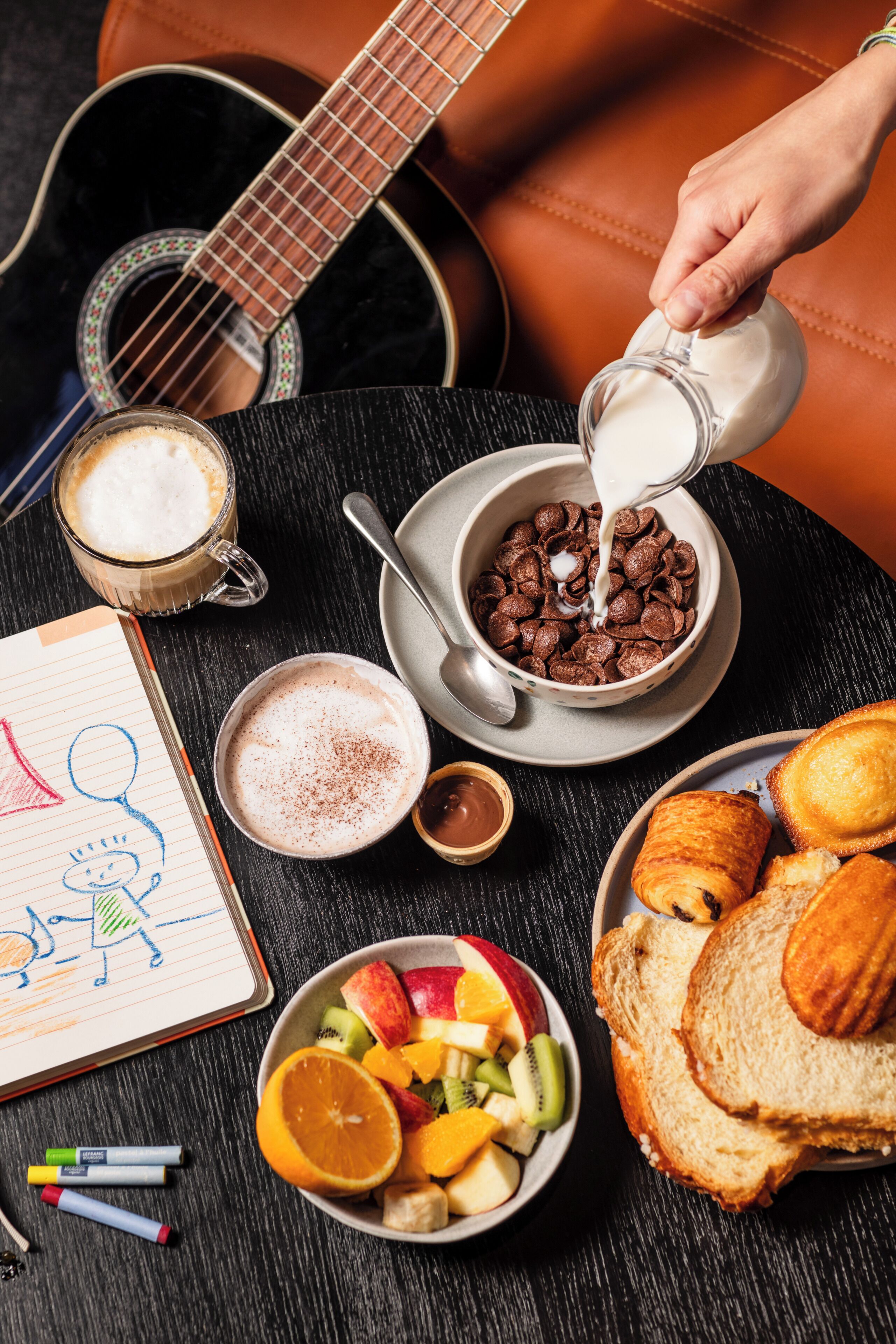 A breakfast setting with a guitar, a cup of coffee, cereal, fruit, and pastries on a table.