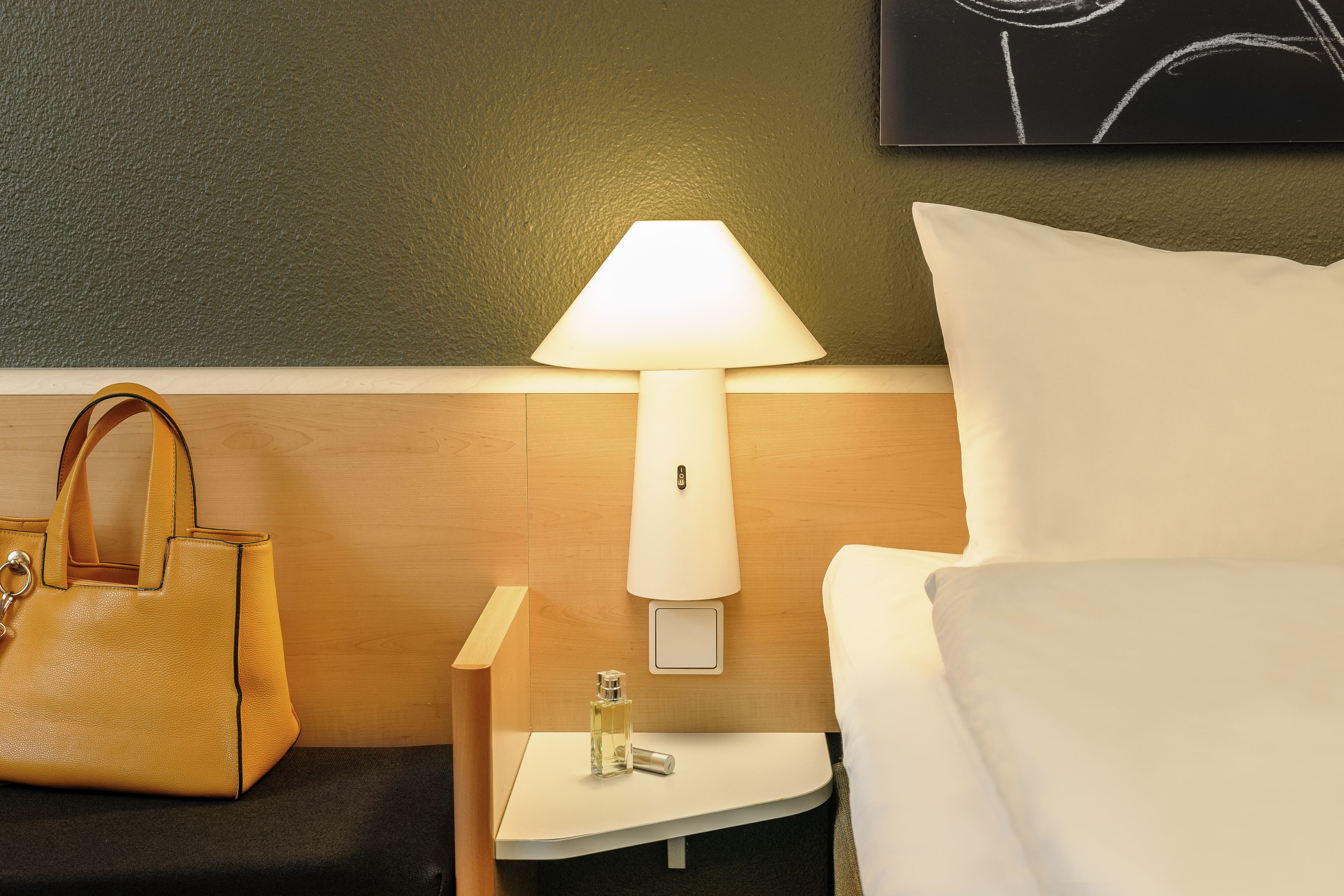 A hotel room with a bed, a nightstand with a lamp, a bottle of perfume, and a handbag.