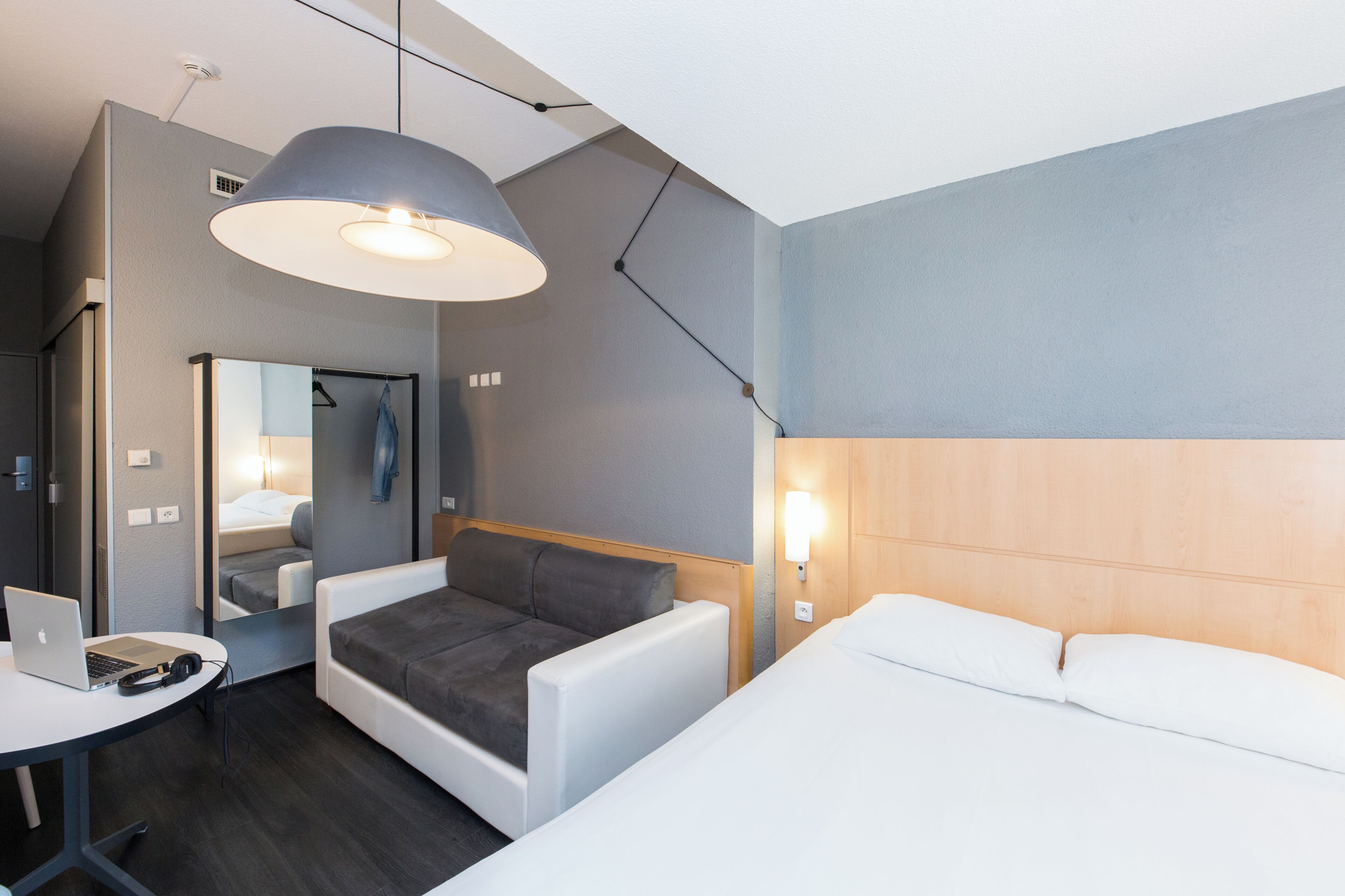 Una habitación de hotel moderna con una cama, un escritorio con una computadora portátil, un sofá, una lámpara colgante y un espejo.