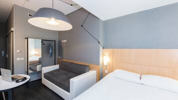 Una habitación de hotel moderna con una cama, un escritorio con una computadora portátil, un sofá, una lámpara colgante y un espejo.