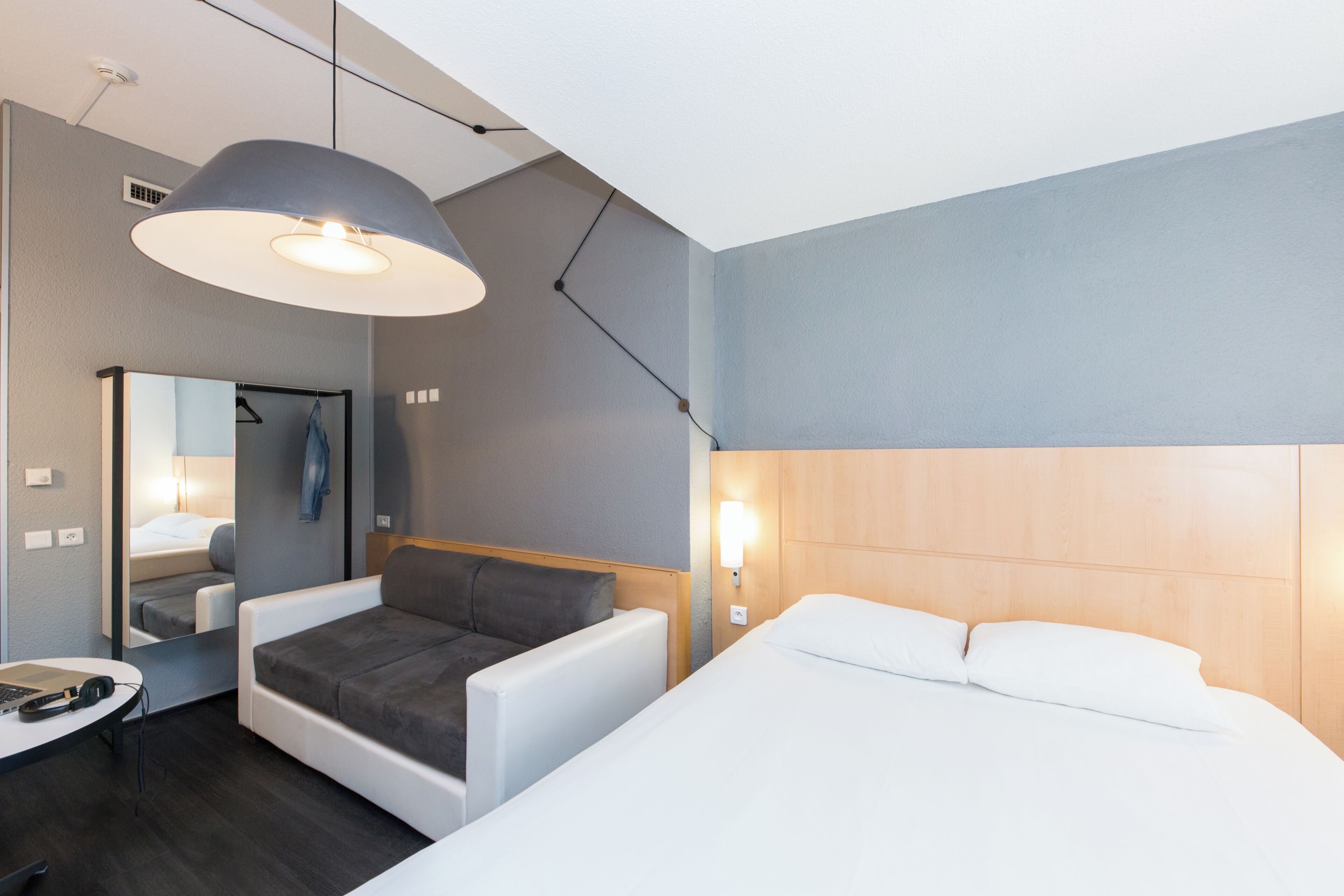 Una habitación de hotel moderna con cama, sofá, escritorio con una computadora portátil, espejo y una lámpara colgante.