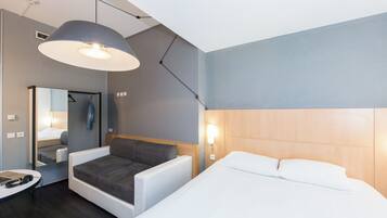 Una habitación de hotel moderna con cama, sofá, escritorio con una computadora portátil, espejo y una lámpara colgante.