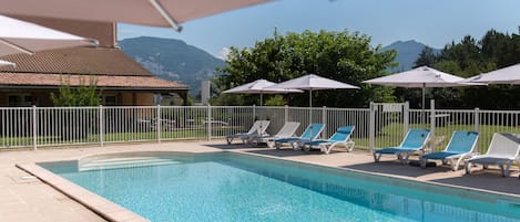 A swimming pool with lounge chairs and umbrellas, surrounded by a fence, with mountains in the background.