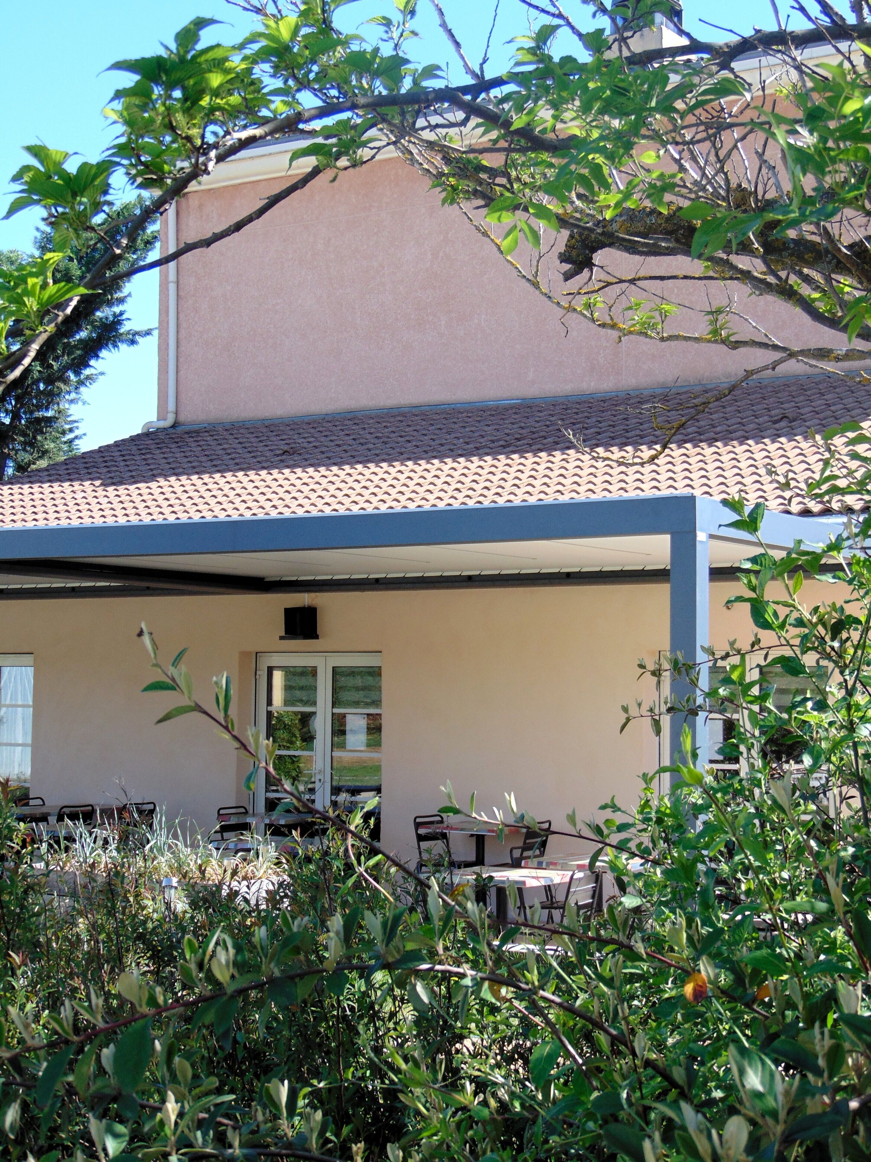 Un espace extérieur abrité, avec des tables et des chaises, une porte, et un bâtiment avec un toit en tuiles en arrière-plan.