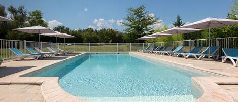 Una piscina rectangular con borde de azulejos, rodeada por una cerca blanca. Hay varias sillas de descanso y sombrillas a lo largo de la piscina.