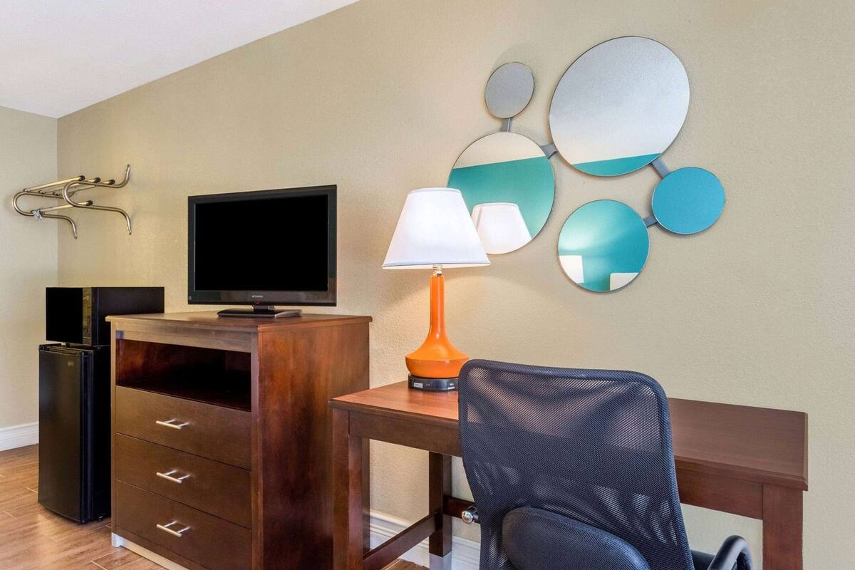 Habitación de hotel con televisión de pantalla plana, un escritorio de madera, una silla, una lámpara y perchero en la pared.