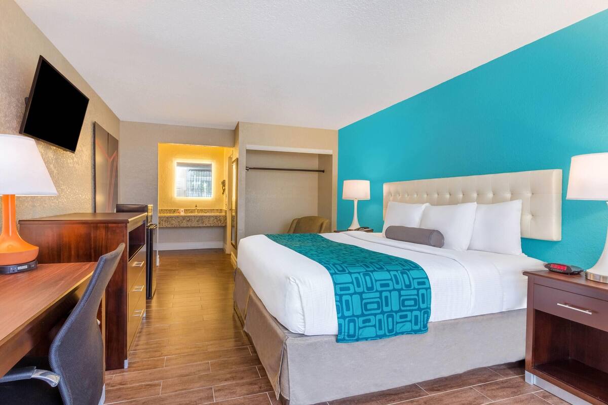 Habitación de hotel con cama, mesitas de noche, escritorio, silla, televisor montado en la pared y baño visible a través de una puerta abierta.
