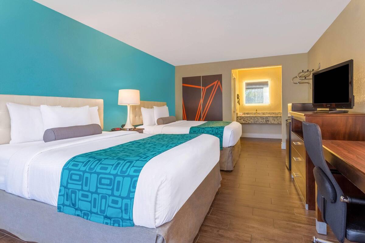 Habitación de hotel con dos camas, un escritorio, un televisor y un baño a la vista a través de una puerta abierta.