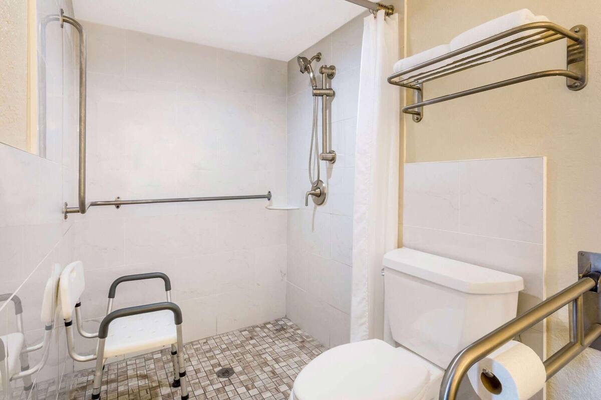 Un baño con ducha, inodoro, barras de apoyo y una silla accesible para sillas de ruedas.