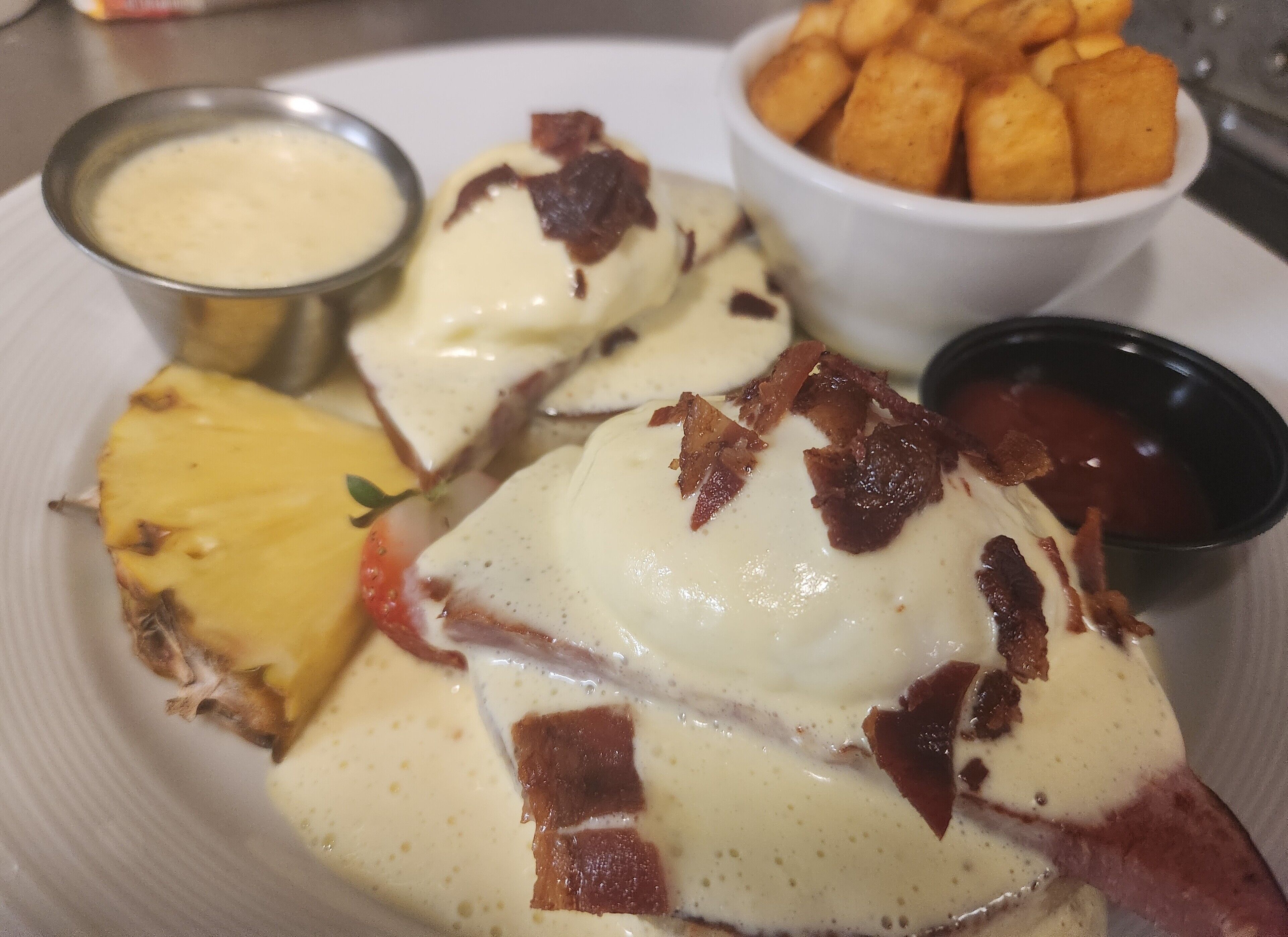 A plate with a slice of pineapple, a piece of ham topped with cheese and bacon, a small bowl of sauce, and a bowl of crispy snacks.