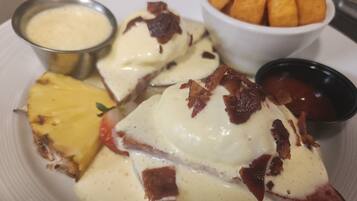 A plate with a slice of pineapple, a piece of ham topped with cheese and bacon, a small bowl of sauce, and a bowl of crispy snacks.