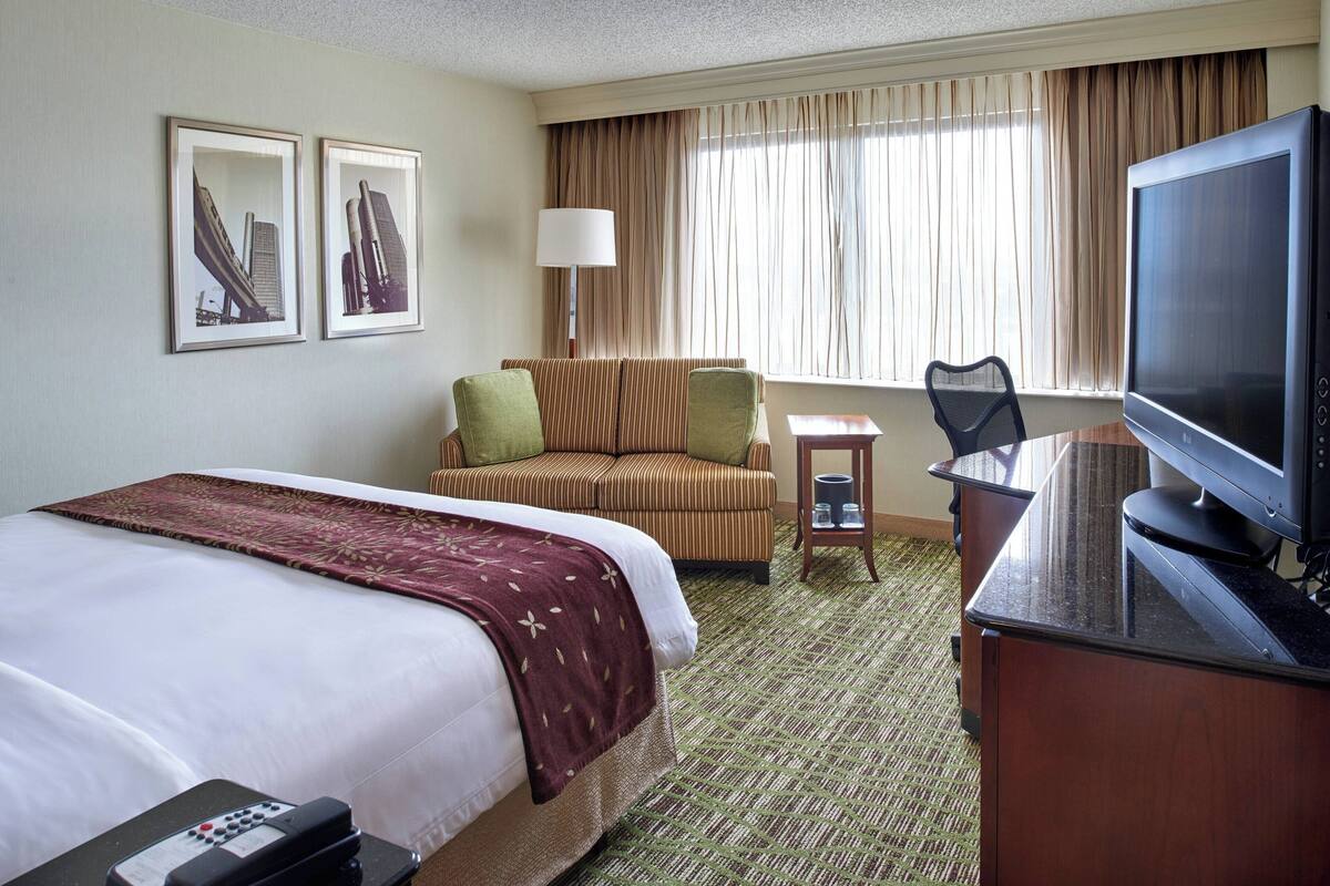 A hotel room with a large bed, a desk with a television, a chair, and a small table. There are curtains on the window and two framed pictures on the wall.