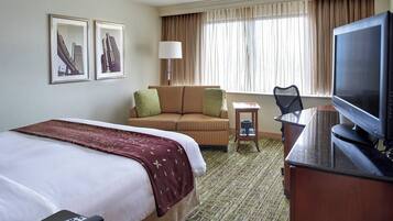 A hotel room with a large bed, a desk with a television, a chair, and a small table. There are curtains on the window and two framed pictures on the wall.