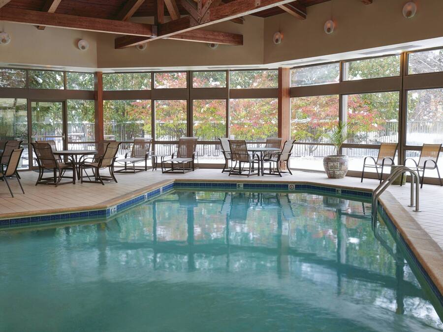 An indoor swimming pool with tiled flooring, wooden deck chairs, and large windows offering views of trees and a fence.