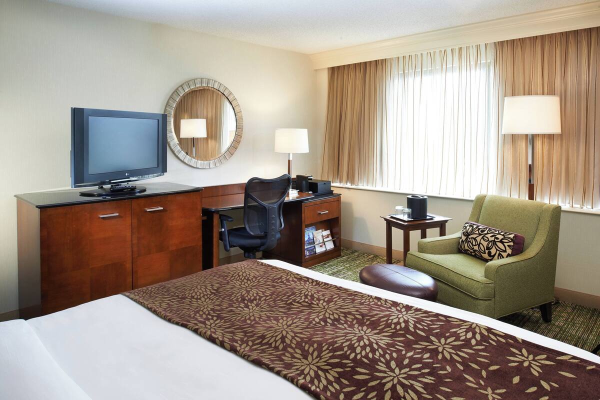 A hotel room with a bed, a desk with a computer, a chair, a television, and a window with curtains.