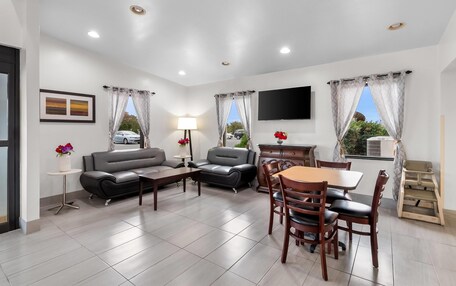 A modern living room with a sofa, armchair, dining table, and chairs. There is a TV mounted on the wall and a small table with a lamp.