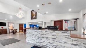 Zona de recepción de hotel con mostrador de piedra, candelabro, escritorio con computadora y área de descanso con sillas y una mesa.