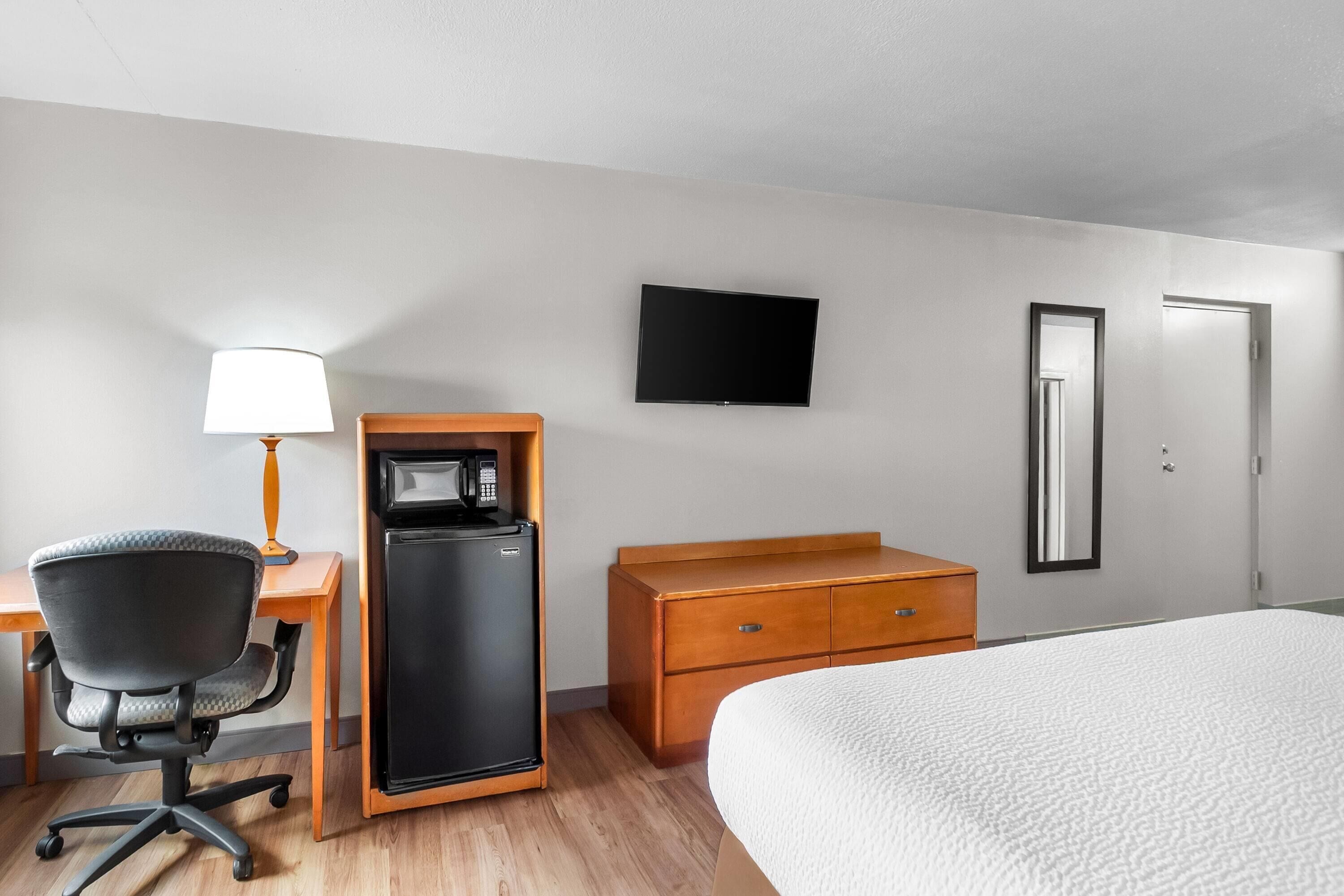 Habitación de hotel con cama, escritorio con lámpara, refrigerador, televisor, cómoda y puerta.