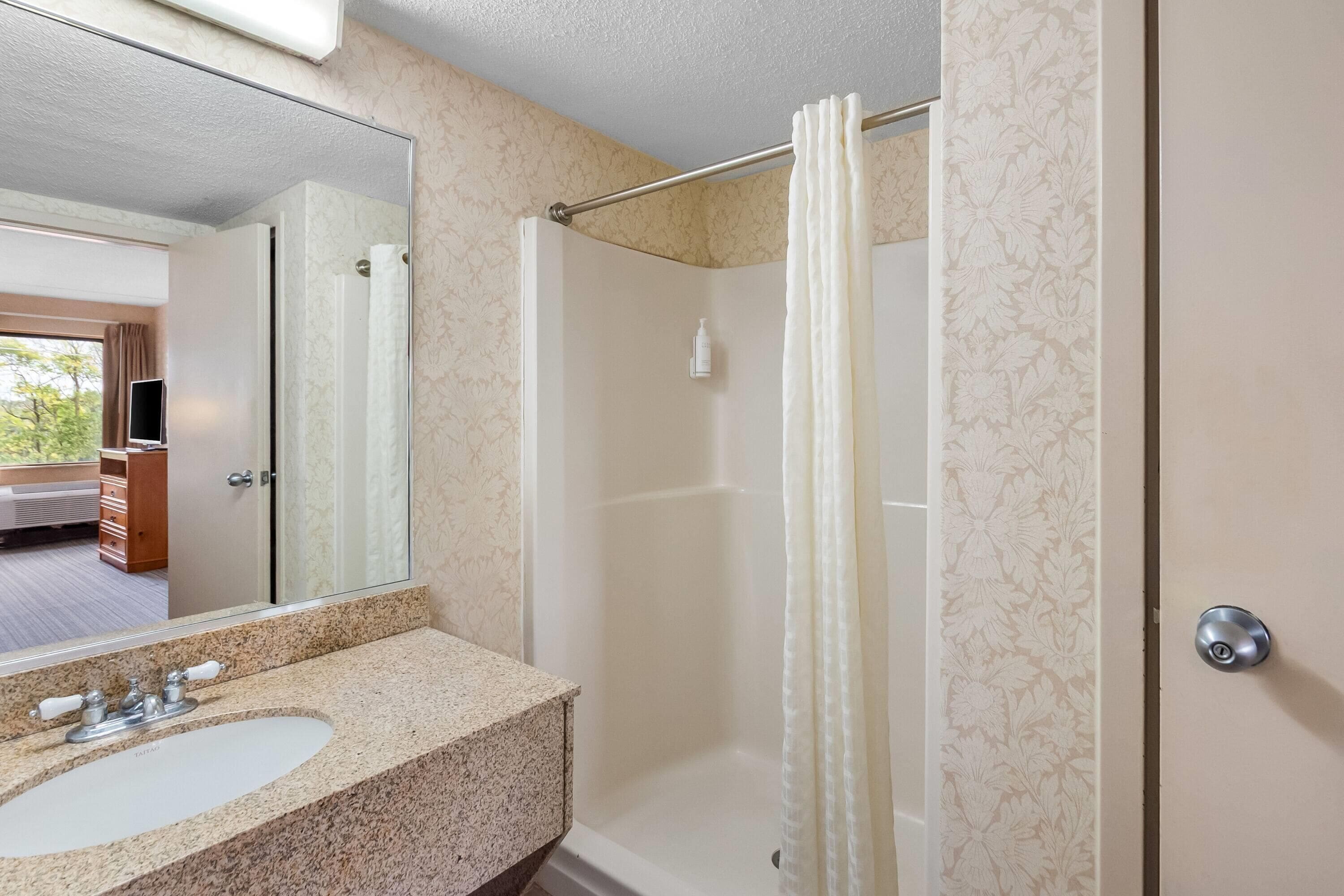 Un baño con lavamanos de encimera de granito, espejo, ducha con cortina y puerta con manija.