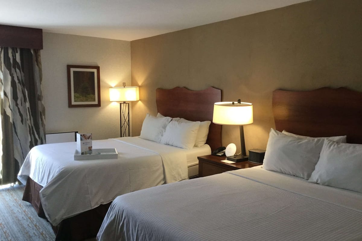 Quarto de hotel com duas camas, cada uma com lençóis brancos, cabeceira, abajures e um quadro na parede.
