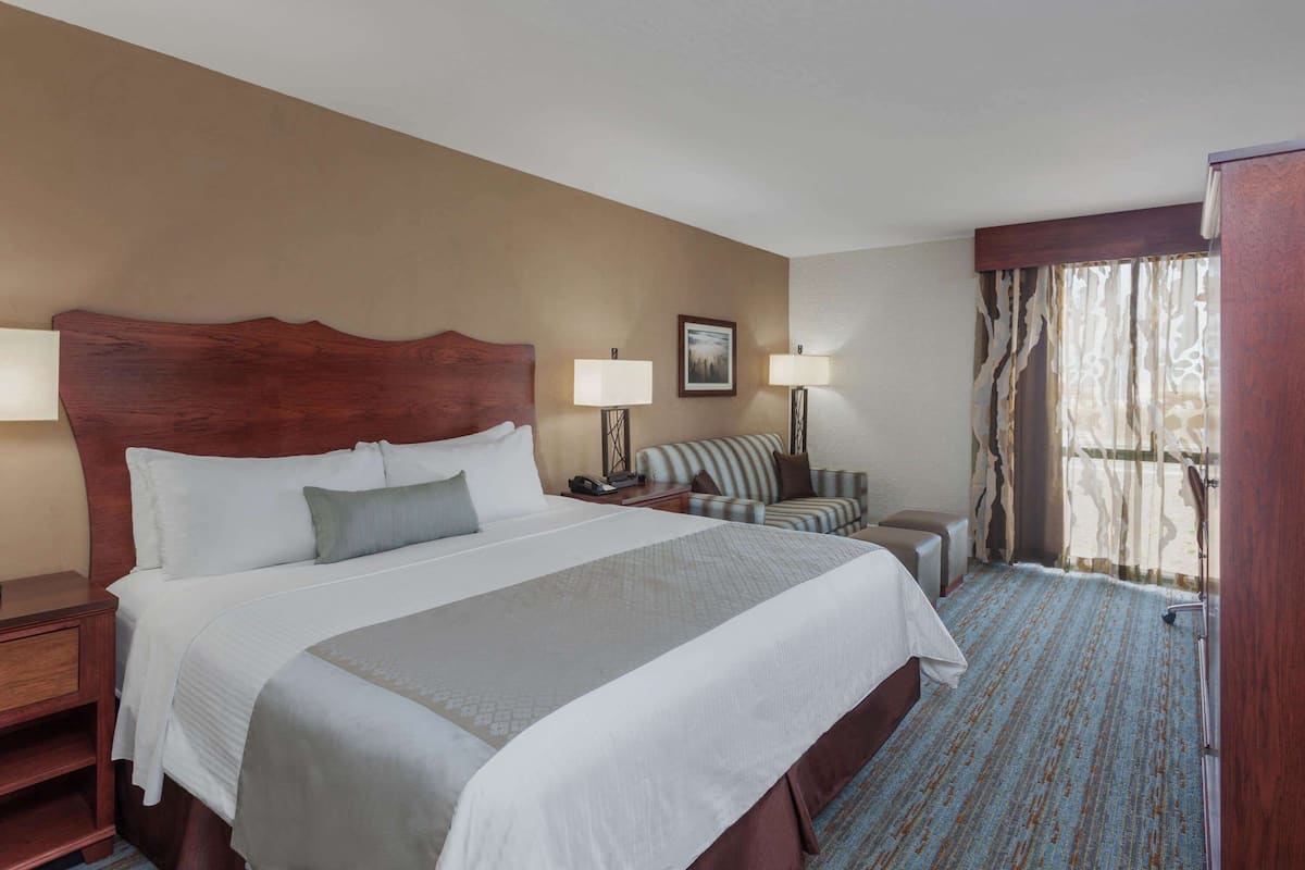 Quarto de hotel com uma cama grande, duas mesas de cabeceira, uma cadeira, uma escrivaninha e uma janela com cortinas.