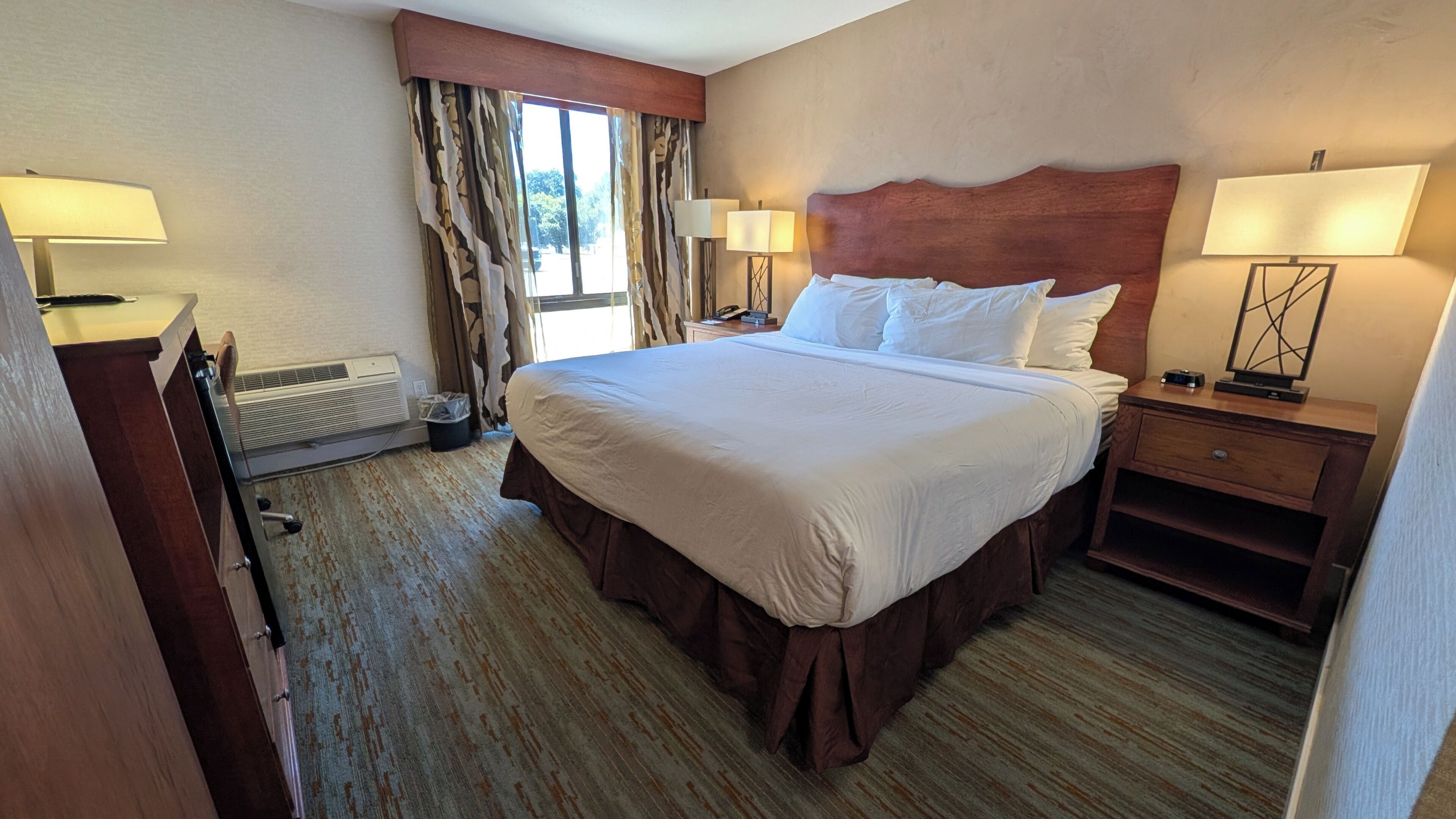 Une chambre d’hôtel avec un grand lit, deux lampes de chevet, une tête de lit en bois, une table de chevet et un radiateur.