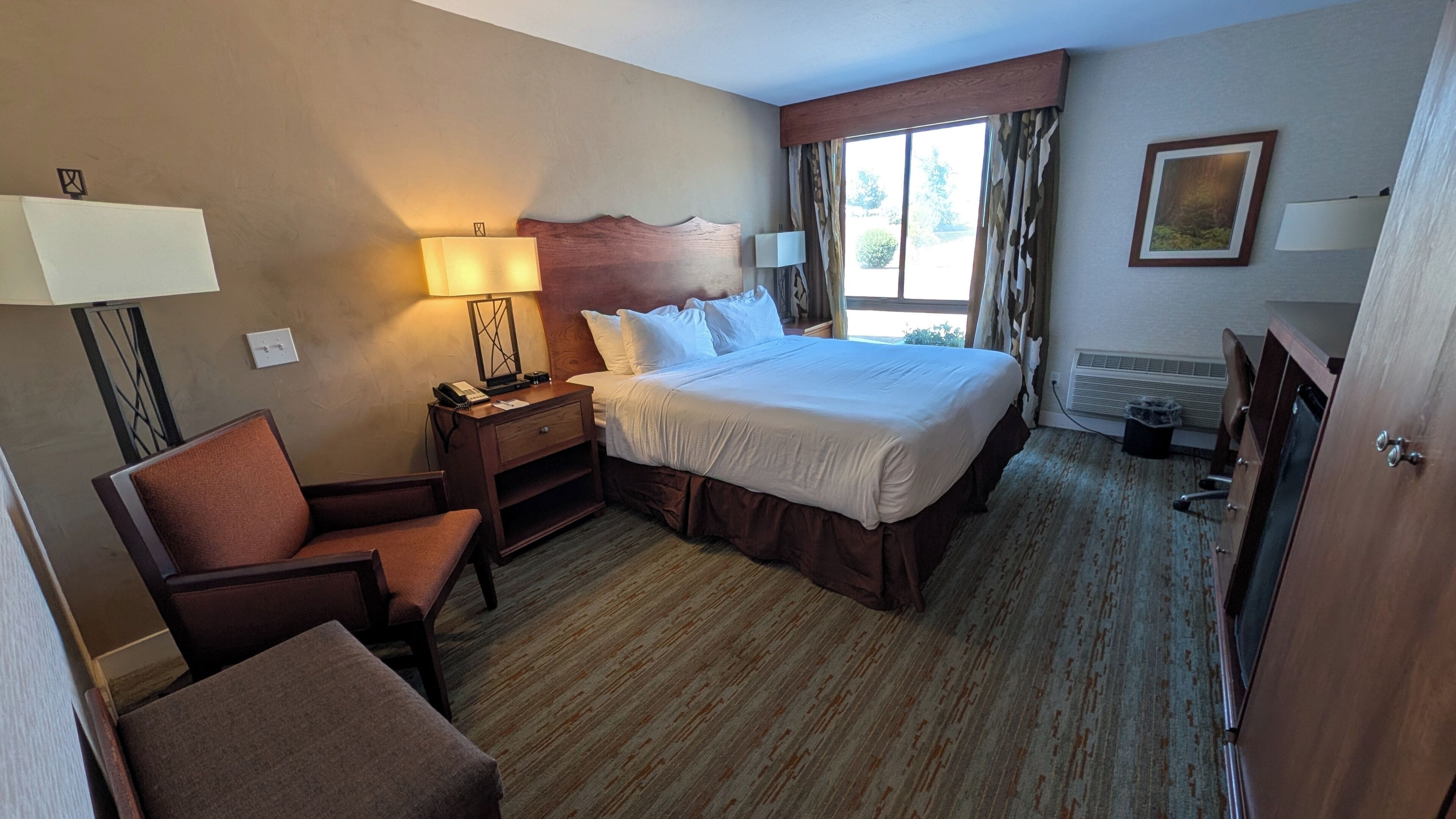 A hotel room with a bed, a desk with a lamp, a chair, a window with curtains, and a painting on the wall.