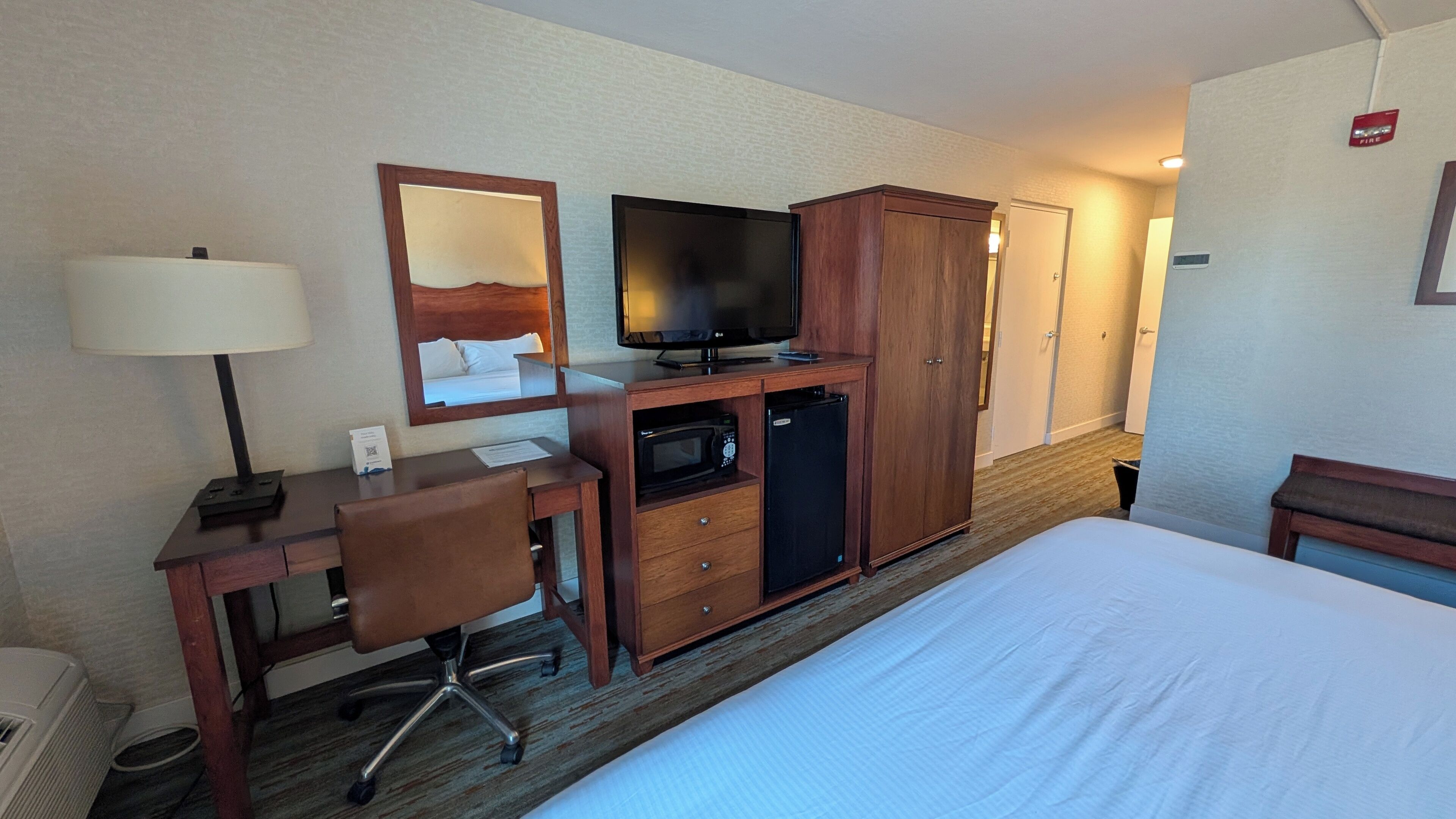 Quarto de hotel com cama, escrivaninha, cadeira, televisão, guarda-roupa e micro-ondas.