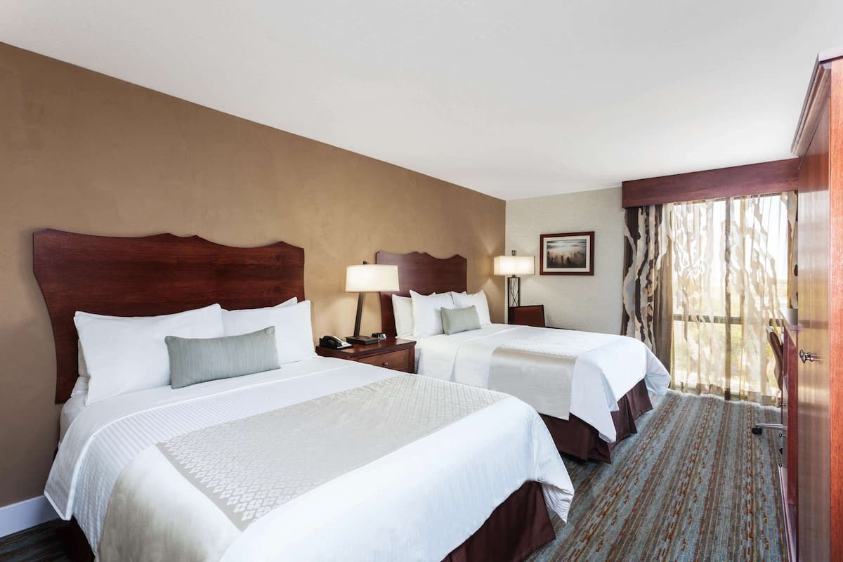 Quarto de hotel com duas camas, cabeceiras de madeira, roupa de cama branca, mesa de cabeceira com abajur, quadro emoldurado na parede e janela com cortinas.