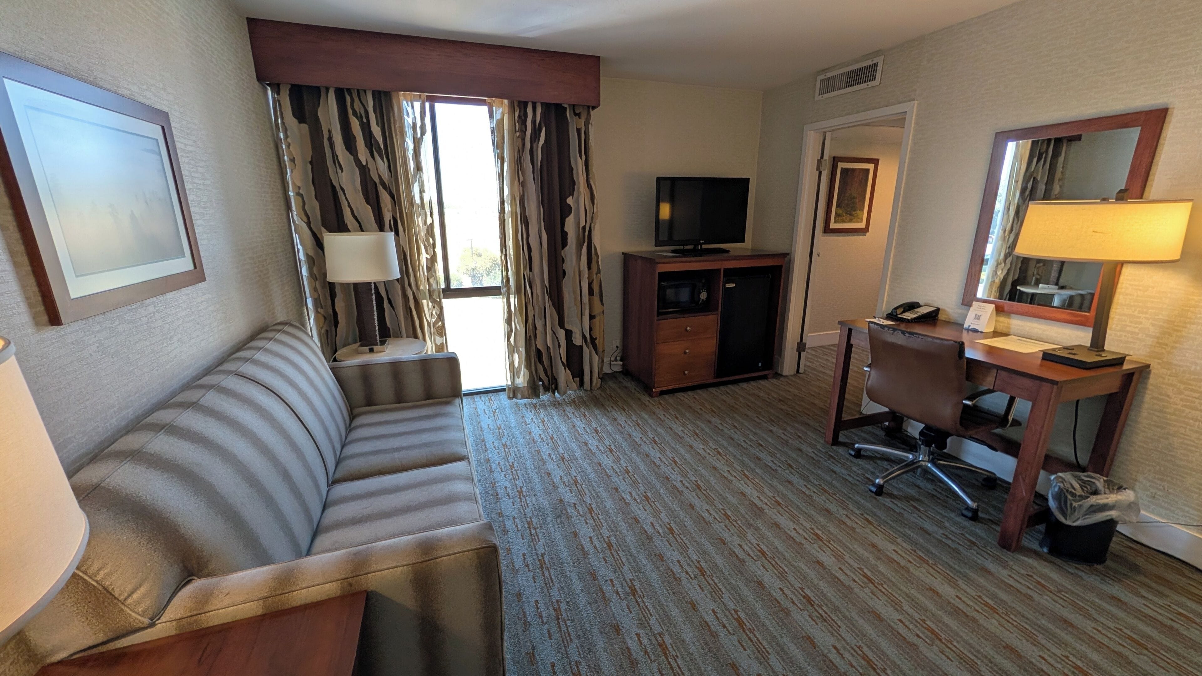 Habitación de hotel con sofá, escritorio con silla, televisión, lámpara y ventana con cortinas.