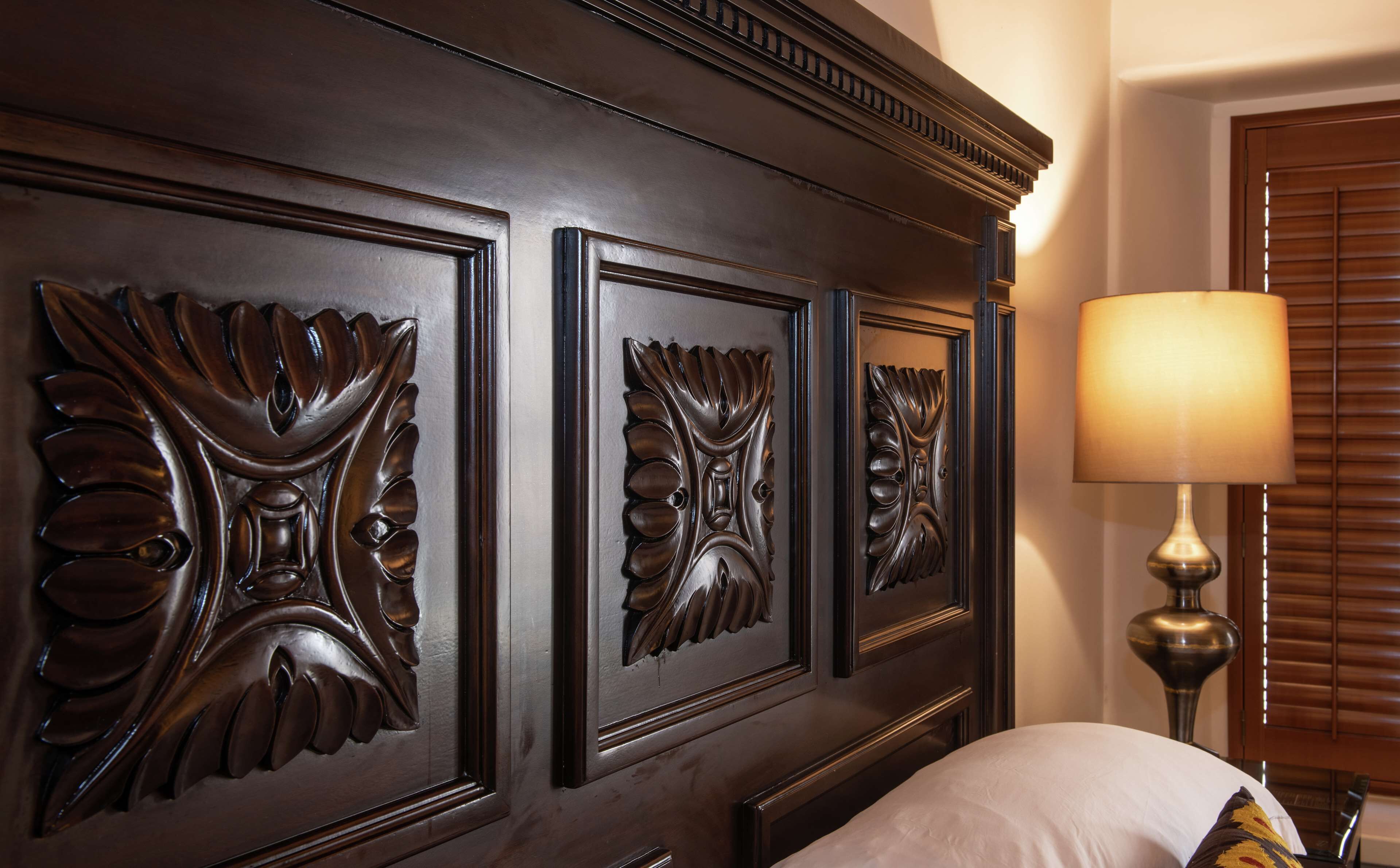 A bedroom with a dark wood headboard featuring ornate leaf carvings, a bedside lamp, and a white pillow.