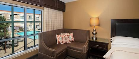 A hotel room with a large window, a brown leather sofa with patterned cushions, a bedside table with a lamp, and a bed with a white comforter.