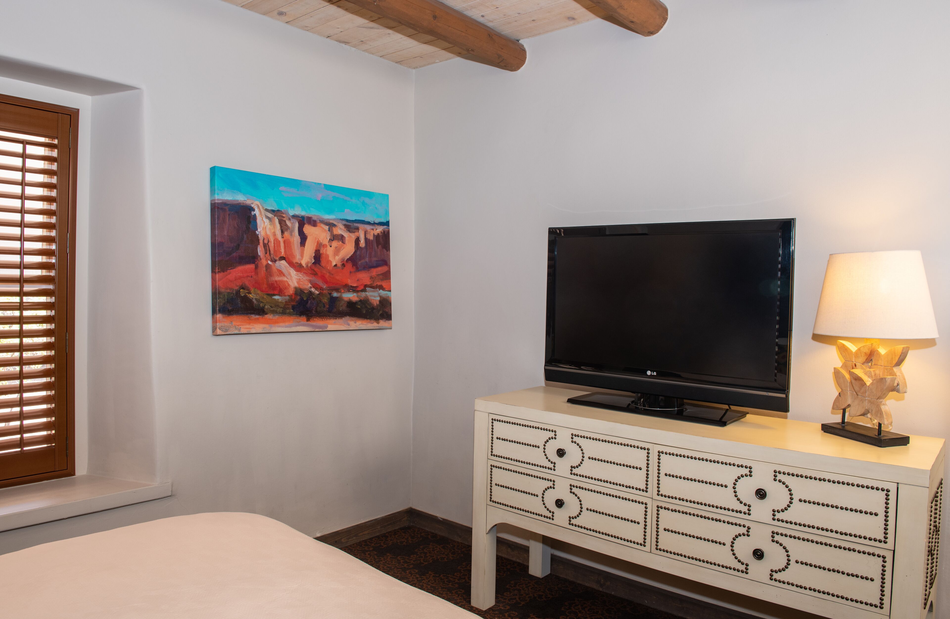 A hotel room with a bed, a television on a white dresser, a painting of a landscape, and a window with blinds.
