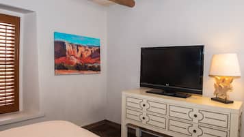 Une chambre d’hôtel avec un lit, un téléviseur posé sur une commode blanche, un tableau représentant un paysage et une fenêtre avec des stores.