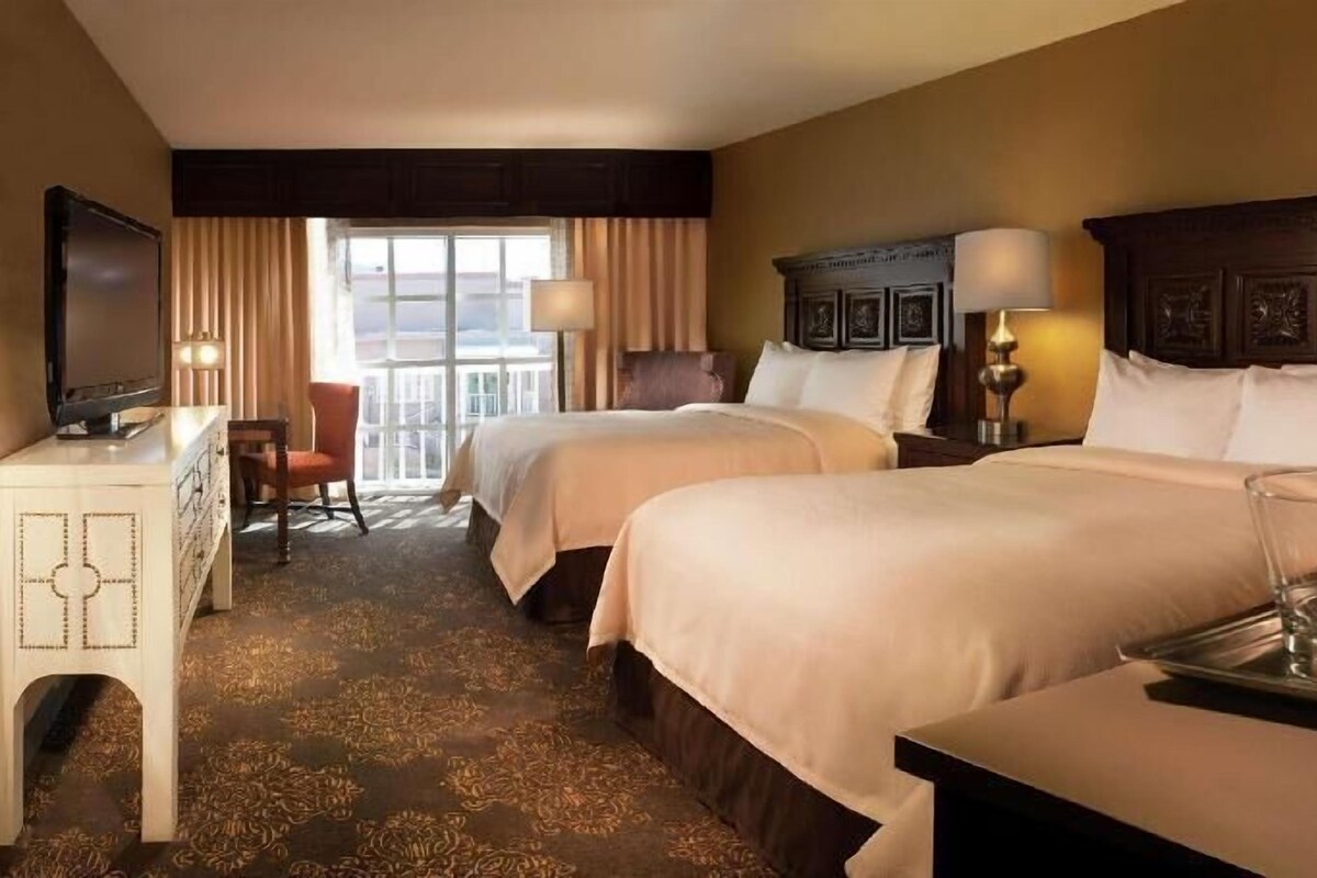 A hotel room with two beds, a desk, a chair, a TV, and a window with curtains.