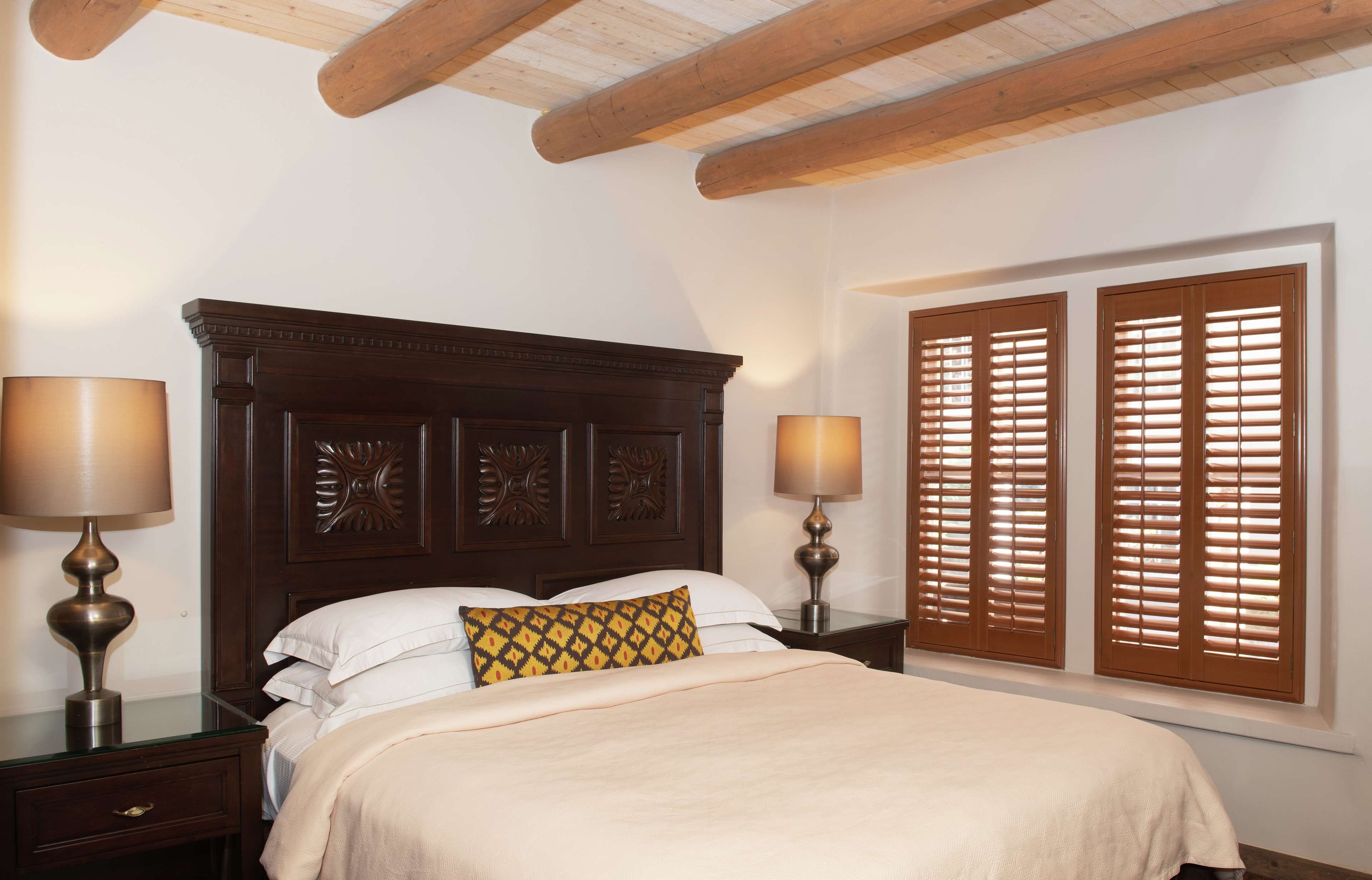 A bedroom with a wooden headboard, two bedside lamps, a window with wooden shutters, and a bed with white bedding and a patterned pillow.