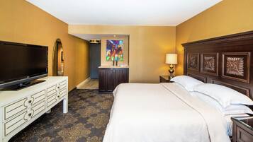 A hotel room with a large flat-screen TV, a bed with white linens, a dresser, a desk with a lamp, and a painting on the wall.