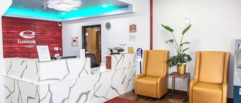 Un espace d’accueil doté d’un comptoir en marbre, de deux fauteuils en cuir, d’une plante en pot et d’un tapis rouge orné du nom de l’hôtel.
